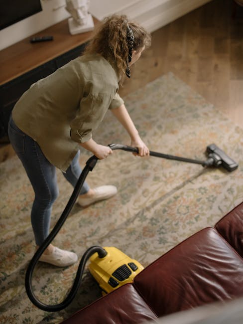 A woman with curly hair, dressed in a beige jacket, dark jeans, and white sneakers, is performing surface cleaning on a patterned area rug using a yellow and black vacuum cleaner. She is standing on a wooden floor in a room with soft lighting, a dark wooden sideboard, and a white vase on top. The rug appears fresh and clean, indicating recent deep cleaning or sanitisation. The scene reflects domestic cleaning efforts aimed at maintaining hygienic and well-maintained living spaces, showcasing the professional cleaning services provided by Haringey Carpet Cleaners, as seen on their website page about upholstery cleaning and rug care in Alexandra Palace, Haringey.