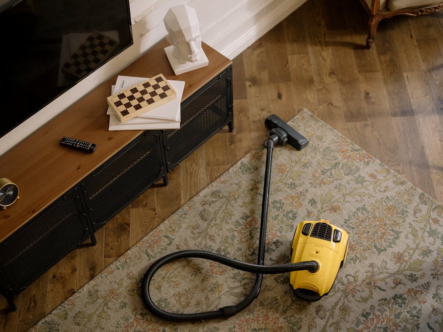 A person kneeling on a patterned carpet in a living room, wearing casual clothing including a beige jacket and jeans, using a yellow and black vacuum cleaner with a transparent dust container to perform surface cleaning. The vacuum cleaner's hose is connected to the main unit, which is positioned on the carpet, and the person appears to be focusing on cleaning the rug. The room features warm lighting, with a wooden floor in the background. The scene emphasizes deep cleaning and maintenance, aligning with the services provided by Haringey Carpet Cleaners, as highlighted on the webpage about avoiding hidden carpet cleaning charges in Haringey.