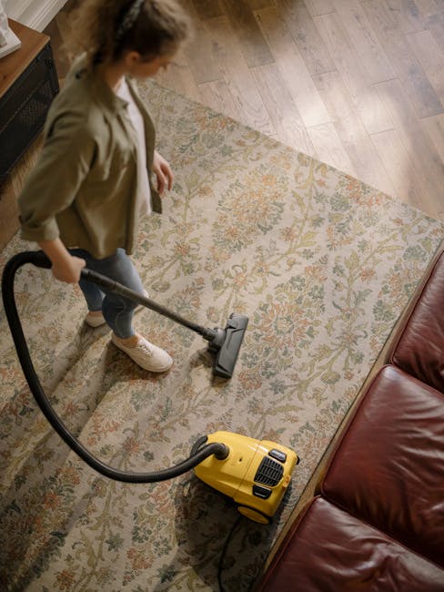 A woman using a yellow vacuum cleaner to perform surface cleaning on a patterned area rug in a living room, with wooden flooring and a leather sofa visible nearby. The room is well-lit, and the rug ap