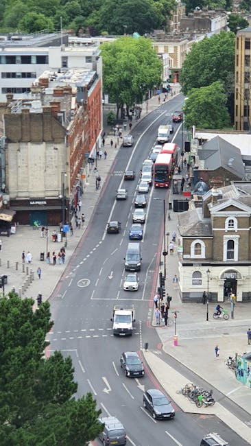 An aerial view of a busy urban street in Wood Green, Haringey, showing a mix of residential and commercial buildings, including a small white church with a grey roof. The street has multiple lanes with a steady flow of vehicles, including cars and buses, and is lined with green trees providing shade. Pedestrians are walking along the sidewalks, some crossing at designated crosswalks. The scene features a variety of building styles, from modern to older brick structures, with clear signage indicating shops and local businesses. Bright daylight illuminates the scene, highlighting the clean and well-maintained surfaces of the roads and sidewalks, reflecting good urban hygiene. This image depicts a typical lively neighborhood atmosphere in Haringey, ideal for residential and commercial activities, with a focus on community and transportation infrastructure, suitable for promoting professional cleaning services like those offered by Haringey Carpet Cleaners on their website for same-day emergency cleaning options in the area.