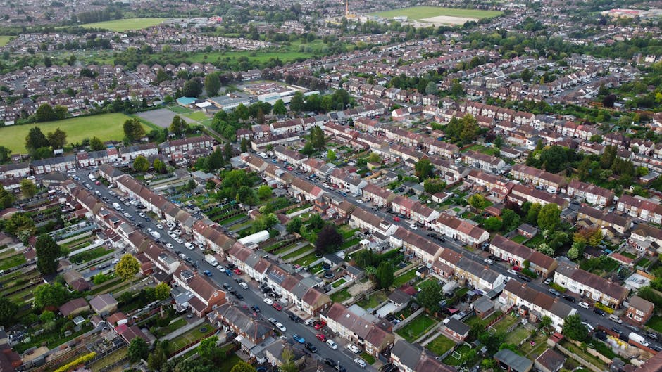 Aerial view of a residential neighborhood in Wood Green, Haringey, featuring rows of terraced houses with front gardens and parked cars lining the streets. The image shows a mix of brick and tiled roofs, lush green trees dispersed throughout the area, and open green spaces in the distance under an overcast sky, illustrating a typical suburban community in North London. The scene is well-lit with natural daylight, providing a clear perspective of the area's layout and housing density, reflecting a peaceful, well-maintained residential environment associated with Haringey Carpet Cleaners' local services.