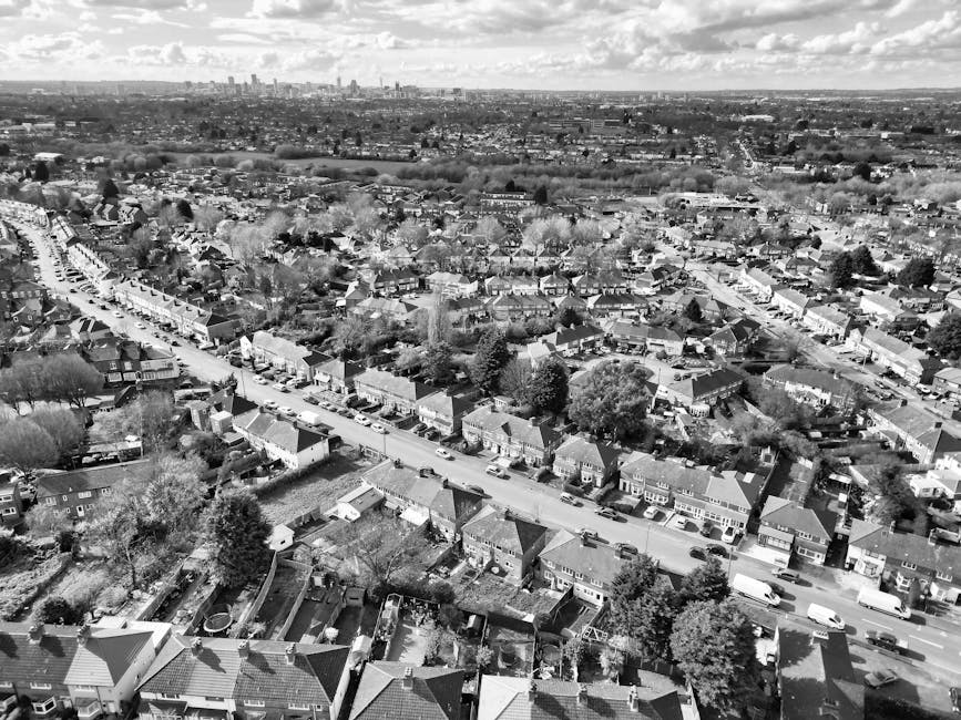 An aerial view of a busy urban street in Wood Green, Haringey, showing a mix of residential and commercial buildings, including a small white church with a grey roof. The street has multiple lanes with a steady flow of vehicles, including cars and buses, and is lined with green trees providing shade. Pedestrians are walking along the sidewalks, some crossing at designated crosswalks. The scene features a variety of building styles, from modern to older brick structures, with clear signage indicating shops and local businesses. Bright daylight illuminates the scene, highlighting the clean and well-maintained surfaces of the roads and sidewalks, reflecting good urban hygiene. This image depicts a typical lively neighborhood atmosphere in Haringey, ideal for residential and commercial activities, with a focus on community and transportation infrastructure, suitable for promoting professional cleaning services like those offered by Haringey Carpet Cleaners on their website for same-day emergency cleaning options in the area.