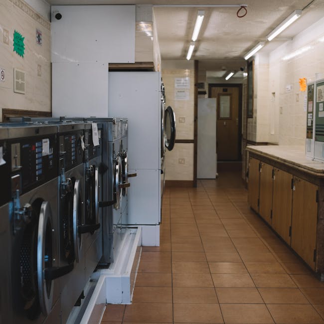 The image depicts a laundry room with stainless steel washing machines on the left side, positioned against a tiled wall. The machines have digital control panels and are arranged in a row, with some doors slightly open. The room features a brown tiled floor and cream-colored, textured wall tiles, with wooden cabinetry along the right side for storage. Overhead, fluorescent lighting illuminates the space evenly. At the back of the room, there is a closed wooden door with a window and visible signage, as well as a white refrigerator or storage unit. The overall appearance suggests a utilitarian environment maintained in a clean and orderly condition. This setting is representative of a commercial laundry or communal cleaning space, which might be serviced or maintained by Haringey Carpet Cleaners, as indicated on the page about access problems for cleaners in Haringey.