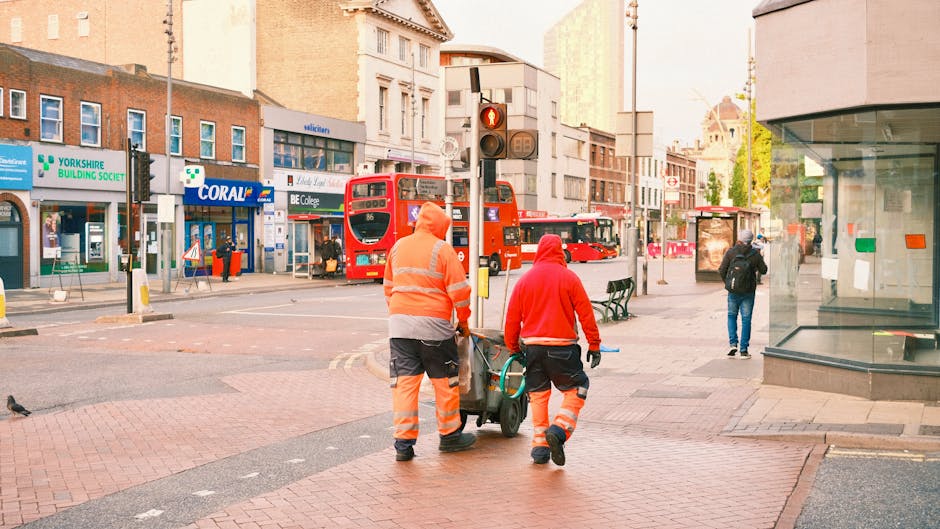 Two street cleaners in high-visibility orange and red uniforms are pushing a cleaning cart along a sidewalk in an urban area of Haringey. The scene features a busy intersection with red double-decker buses stopped at a traffic light, and various shops and buildings on the surrounding streets. The surface of the sidewalk appears clean and well-maintained, with brick paving and smooth concrete sections, under natural daylight. The background includes a mix of commercial establishments, including a pharmacy and a retail store, with pedestrians walking nearby. This image highlights the importance of surface cleaning and maintenance in public spaces, consistent with the services offered by Haringey Carpet Cleaners and other professional cleaning companies.